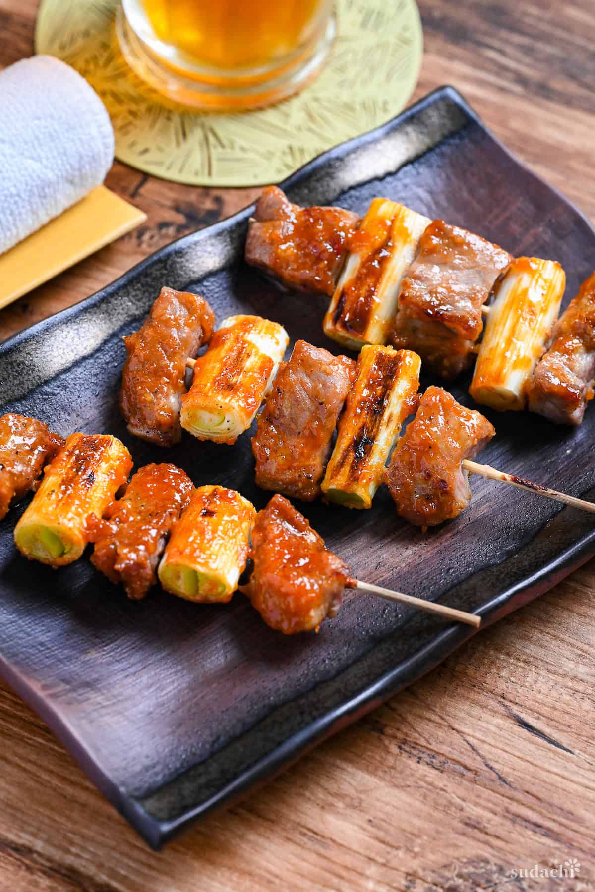 Three Yakiton skewers made with grilled pork shoulder and Japanese leek served on a skewer and brushed with a miso tare (sauce) on a brown rectangular plate
