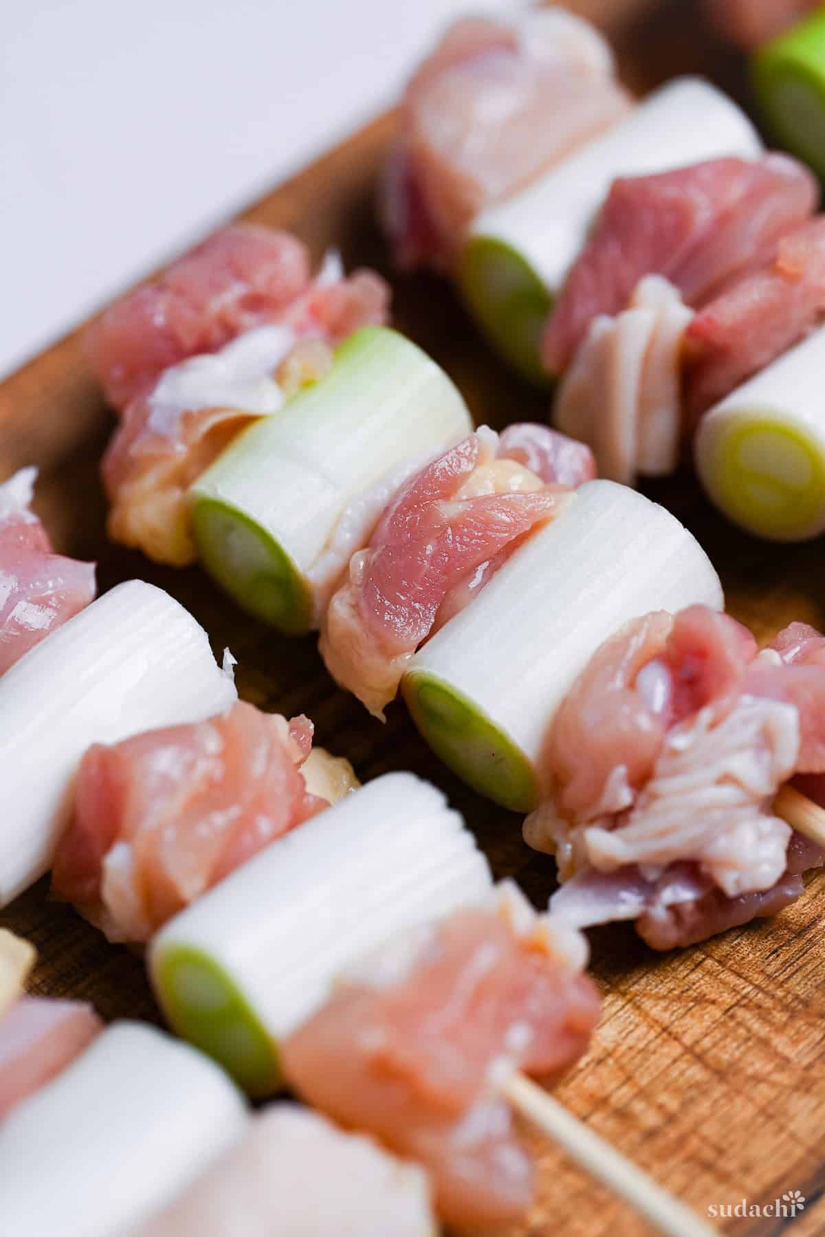 close up of raw negima yakitori (Japanese chicken and leek skewers) threaded onto bamboo skewers and lined up on a wooden chopping board closeup