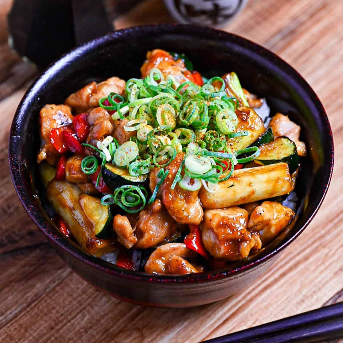 Yakitori donburi made with chicken, zucchini, green onions and bell peppers cooked in a yakitori-style tare (sauce) served in a black bowl and topped with green onion and shichimi