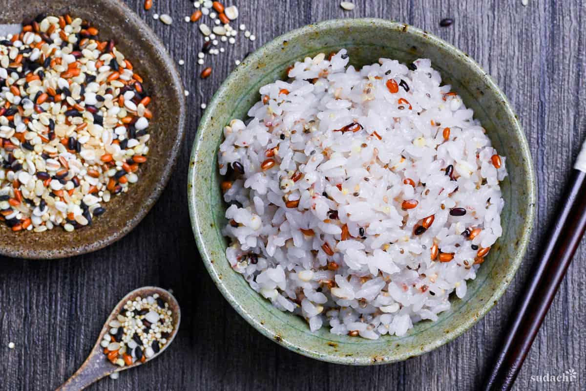 Japanese multigrain rice (zakkokumai) in a green and brown ceramic rice bowl surrounded by various grains and seeds