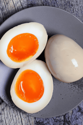 Two Ramen Eggs (Ajitsuke Tamago) with one cut in half to reveal jelly yolk on a gray plate next to wooden chopsticks on a ash-wood background