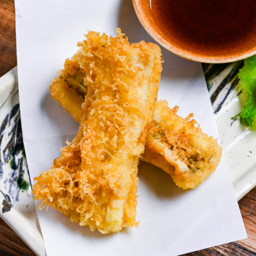 two pieces of conger eel tempura on a rectangular plate with tempura dipping sauce and grated daikon on a perilla leaf