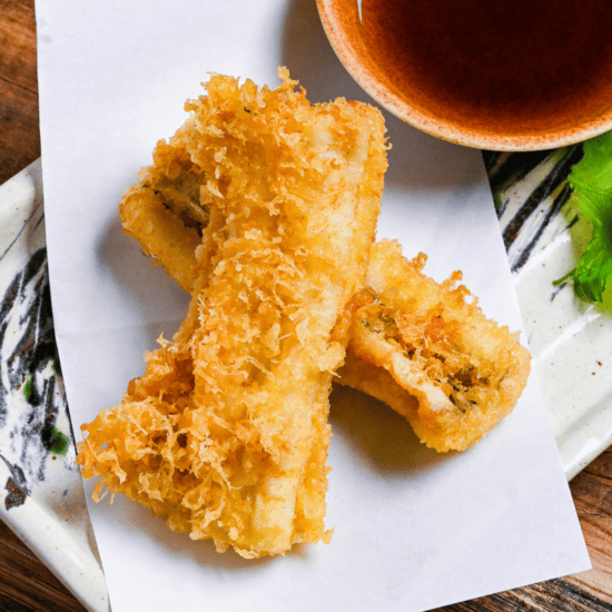 two pieces of conger eel tempura on a rectangular plate with tempura dipping sauce and grated daikon on a perilla leaf
