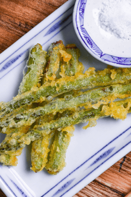 Six pieces of asparagus tempura stacked on a white plate with blue border, with a small round dish of salt next to it and a black pepper grinder in the background