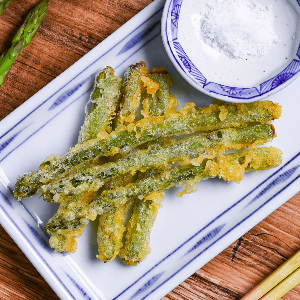 Six pieces of asparagus tempura stacked on a white plate with blue border, with a small round dish of salt next to it and a black pepper grinder in the background