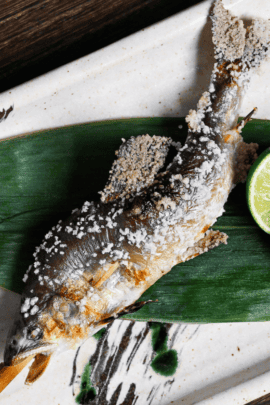 Ayu no Shioyaki (Salt Grilled Sweet Fish) on a bamboo skewer on a leaf with a halved sudachi on a rectangular ceramic plate with black and green brushstroke design