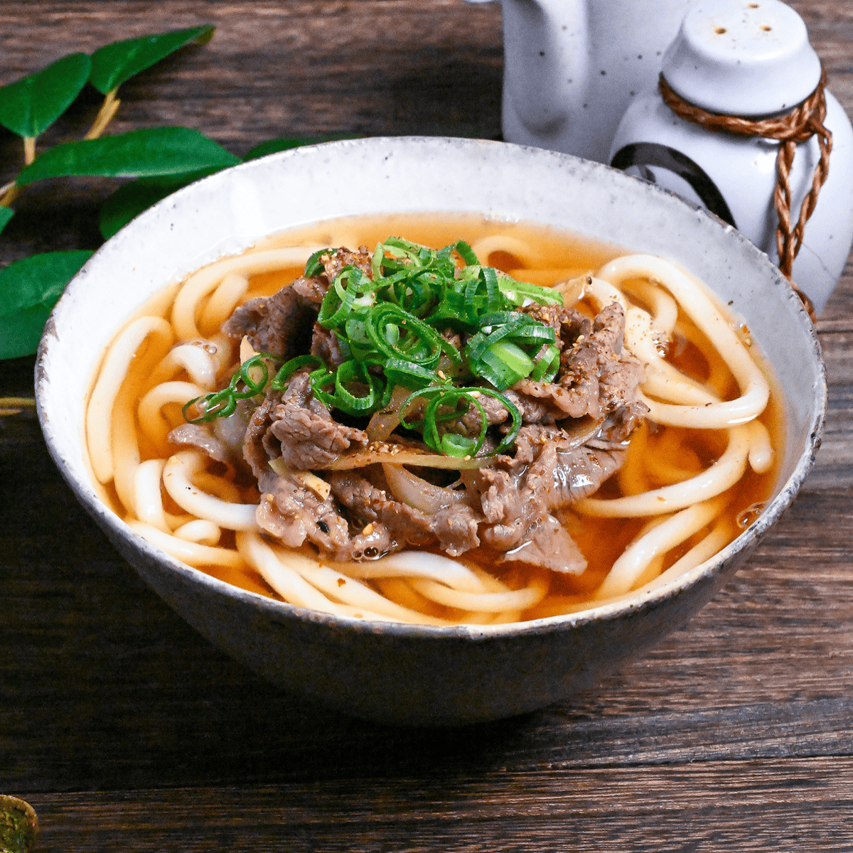 Beef niku udon topped with green onions in a white and gray mottled ceramic bowl on a dark wooden table side view