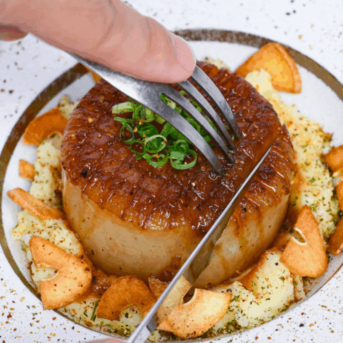 Cutting into a daikon steak served over mashed potato and sprinkled with crispy garlic chips on a white plate with gold rim