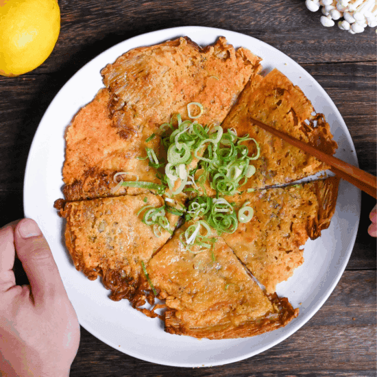 Crispy enoki cheese cut into triangles and served on a white plate with chopped green onions and a small bowl of ketchup on the side. One hand is holding the plate and the other is holding up one piece of enoki cheese with wooden chopsticks.