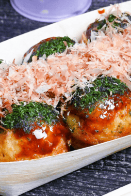 8 crispy gindako style takoyaki topped with glossy brown sauce, aonori and bonito flakes on a black wood-effect background