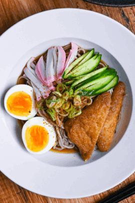 Hiyashi kitsune soba in a white dish topped with marinated tofu pouches, cucumber, chopped green onion, kamaboko fish cakes and boiled eggs on a wooden background top down