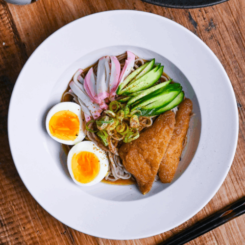 Hiyashi kitsune soba in a white dish topped with marinated tofu pouches, cucumber, chopped green onion, kamaboko fish cakes and boiled eggs on a wooden background top down