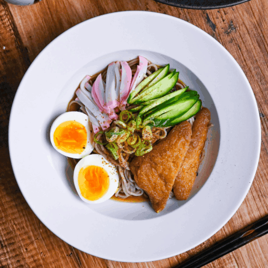 Hiyashi kitsune soba in a white dish topped with marinated tofu pouches, cucumber, chopped green onion, kamaboko fish cakes and boiled eggs on a wooden background top down