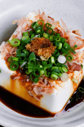Hiyayakko, a block of silken tofu topped with soy sauce, bonito flakes, green onions and grated ginger in a white ceramic dish on a wooden background