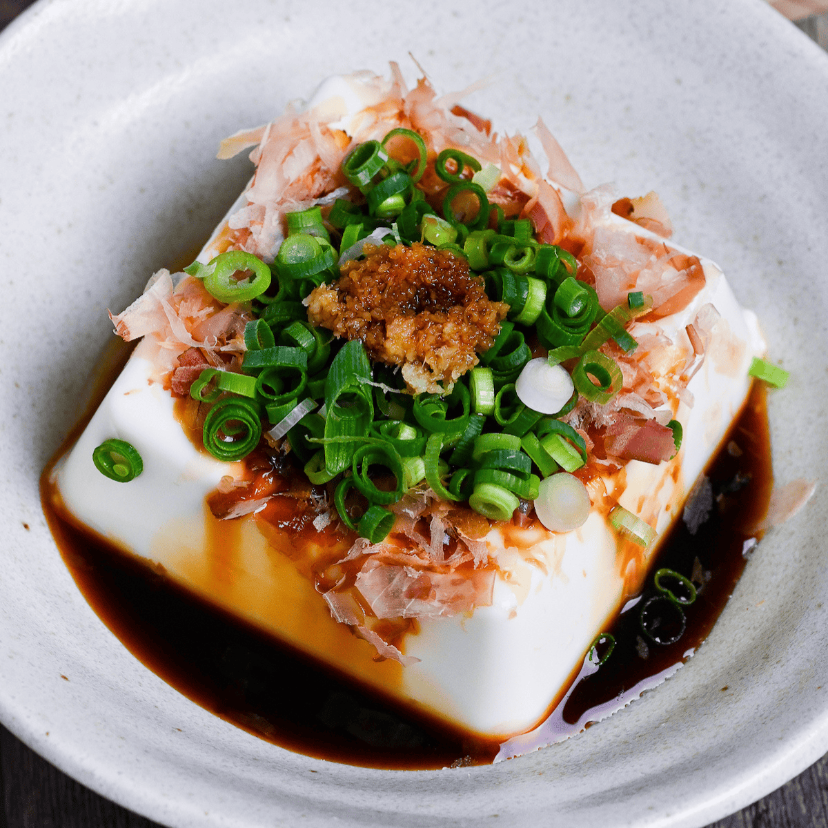 Hiyayakko, a block of silken tofu topped with soy sauce, bonito flakes, green onions and grated ginger in a white ceramic dish on a wooden background