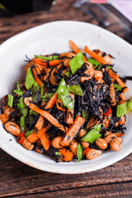hijiki salad in a white dish next to a bottle of soy sauce, dried hijiki and wooden chopsticks on a bamboo shaped rest
