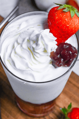 Strawberry mochi milk in a tall glass topped with whipped cream, a strawberry and boiled adzuki beans on a wooden chopping board next to ichigo daifuku on a gray plate