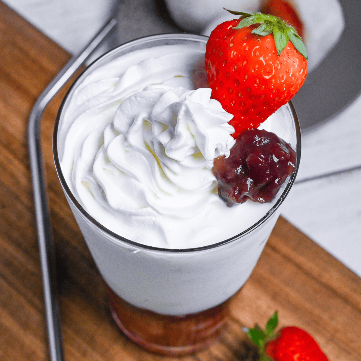 Strawberry mochi milk in a tall glass topped with whipped cream, a strawberry and boiled adzuki beans on a wooden chopping board next to ichigo daifuku on a gray plate