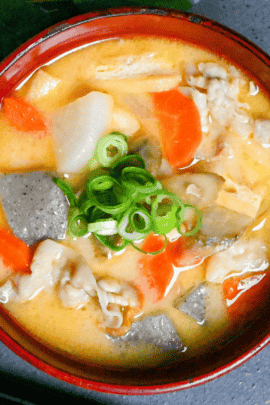 Sake lees soup (kasujiru) made with pork, vegetables and konnyaku in a red bowl on a gray ceramic placemat