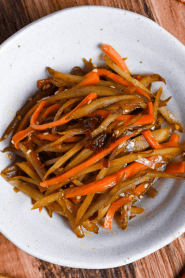 Kinpira gobo made with carrots and burdock root topped with red chili in a white bowl on a wooden background surrounded by dry red chilis