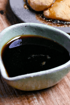 Homemade kuromitsu (Japanese dark sugar syrup) in a small green ceramic jug with it drizzled over mochi in the background