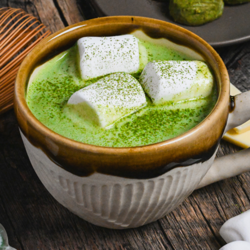 Matcha white hot chocolate in a brown and cream Japanese ceramic mug topped with marshmallows and matcha powder on a dark wooden background