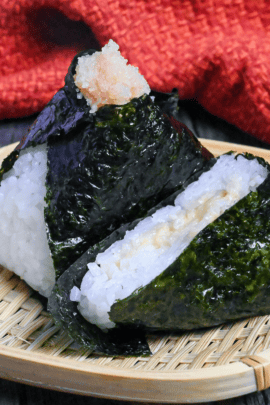 Mentaiko onigiri with one cut in half to show the filling on a small bamboo tray on a dark gray wood-effect background with red fabric in the background