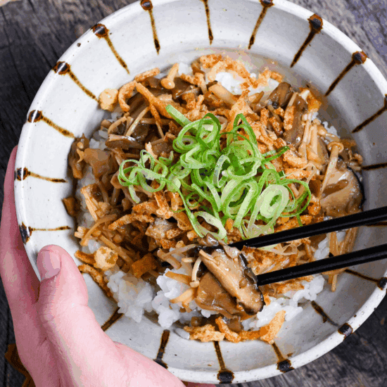 a hand holding mushroom donburi with black chopsticks in the other hand holding up mushrooms and rice