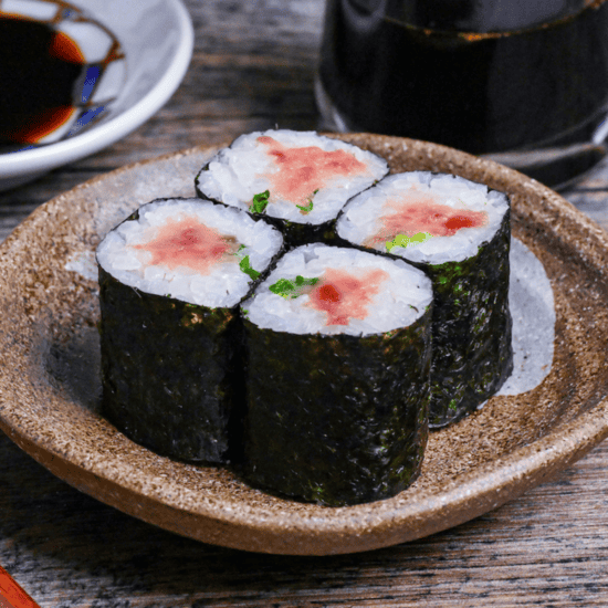 Four pieces of homemade negitoro sushi rolls on a brown serving dish with brush design next to a checkered dipping bowl and glass bottle of soy sauce
