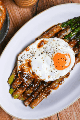 Nikumaki (pork belly wrapped) asparagus topped with sunny side up egg and sprinkled with freshly ground black pepper on a white plate on a wood-effect background