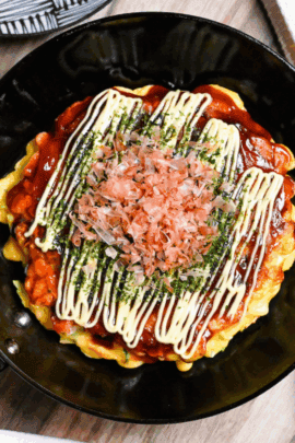 Osaka style okonomiyaki topped with homemade okonomi sauce, mayonnaise, aonori and bonito flakes in a small frying pan with sauce and a plate in the background