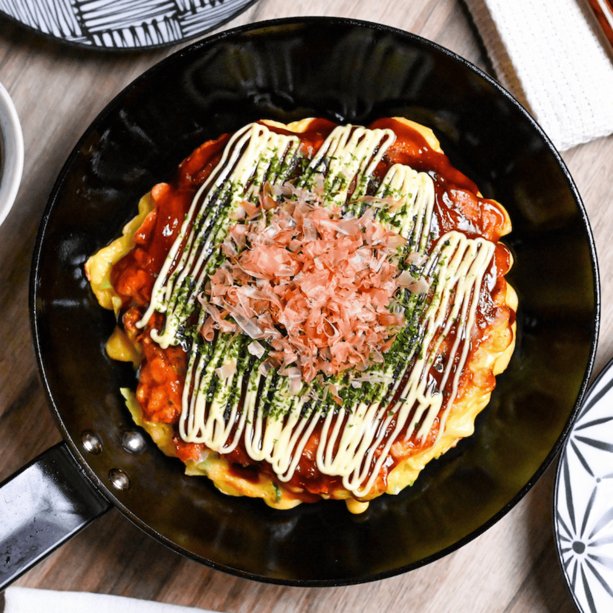 Osaka style okonomiyaki topped with homemade okonomi sauce, mayonnaise, aonori and bonito flakes in a small frying pan with sauce and a plate in the background