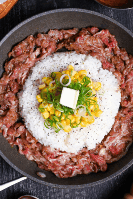 Beef pepper rice in a frying pan on a black wood effect background surrounded by a block of butter, a bowl of sauce and a wooden pepper grinder