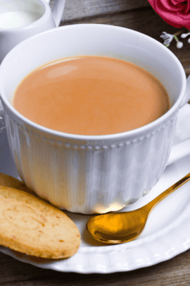 Japanese royal milk tea in a white cup with saucer, shortbread cookies and a golden teaspoon on a light wooden background with sugar cubes, a milk jug and flowers in the background