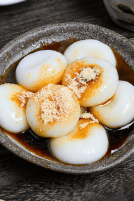 Shiratama dango (mochi balls) topped with kuromitsu (brown sugar syrup) and kinako (soybean powder) in a brown dish on a wooden table