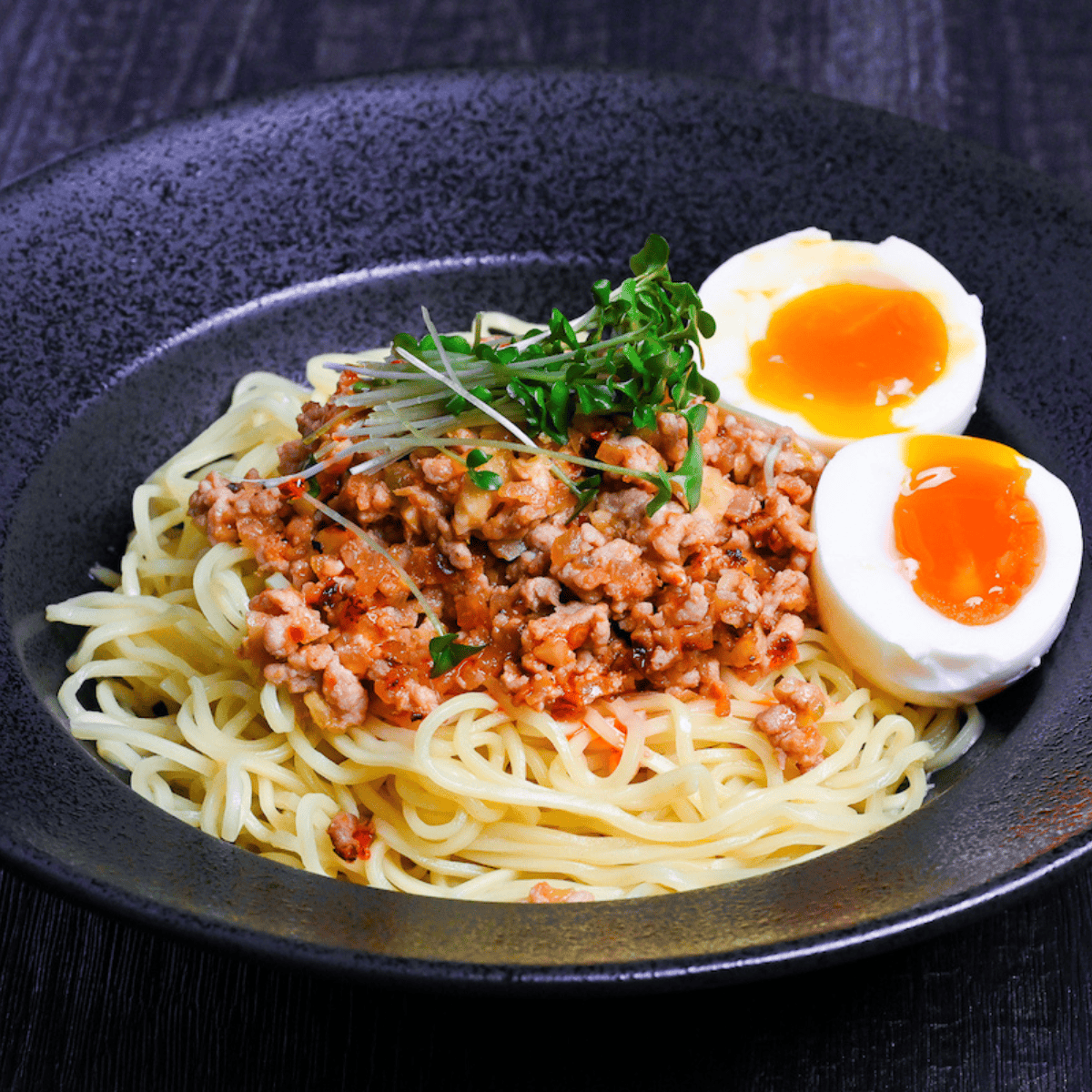 Shirunashi tantanmen topped with soft boiled eggs and broccoli sprouts served in a black dish on a black wood effect background