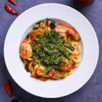 Spicy seafood yaki udon sprinkled with aonori and sesame seeds in a white dish on a gray surface surrounded by dry chilis and a jar of chili oil