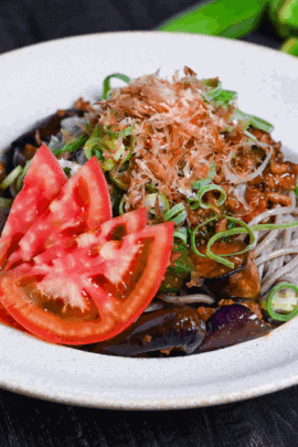 Soba noodle salad made with summer vegetables and miso meat sauce in an off-white bowl topped with chopped green onions, bonito flakes and sliced tomato on a black surface