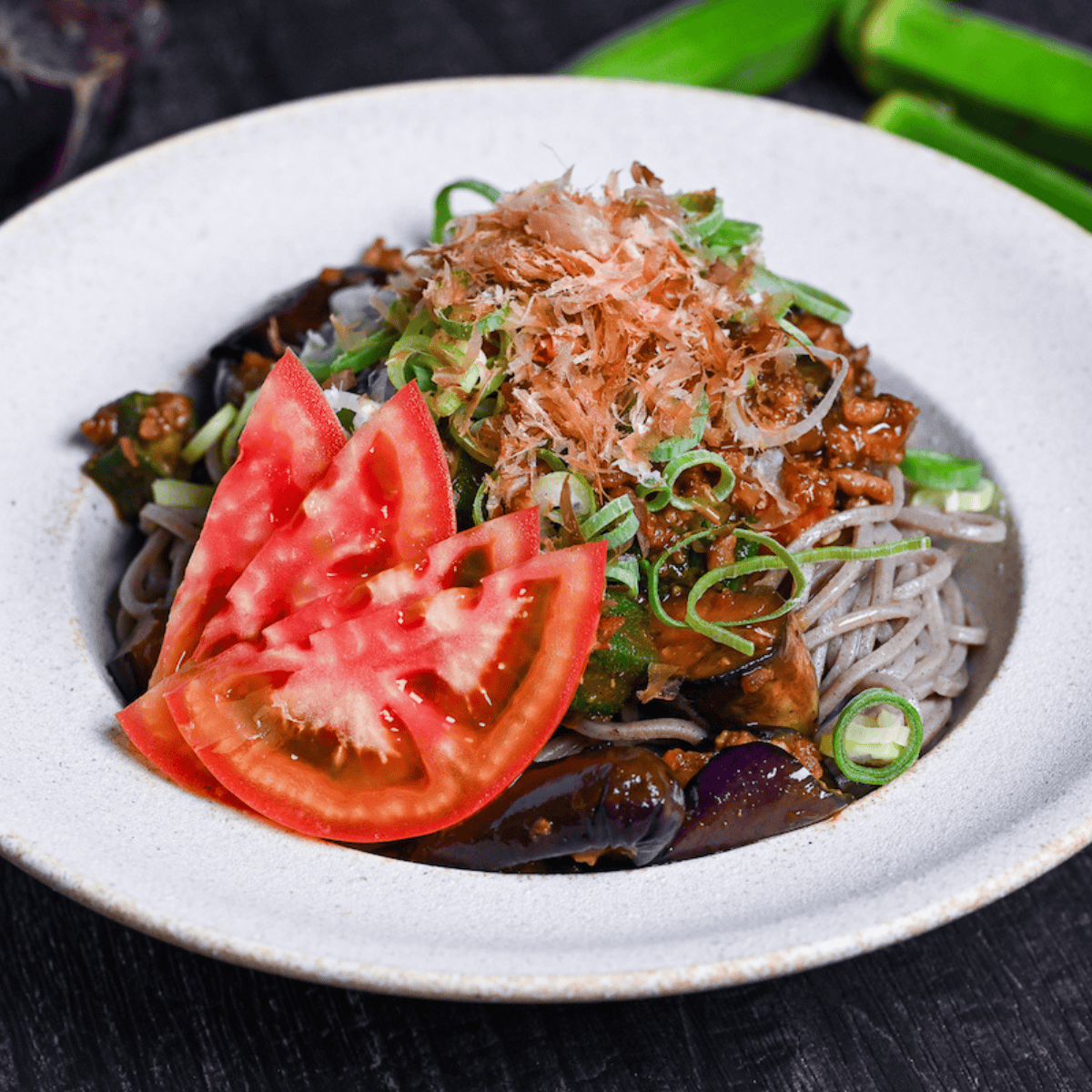 Soba noodle salad made with summer vegetables and miso meat sauce in an off-white bowl topped with chopped green onions, bonito flakes and sliced tomato on a black surface