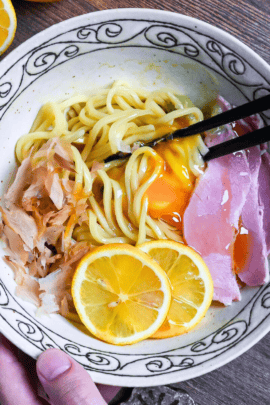 TKM (Tamago Kake Men) topped with bonito flakes, lemon slices, ham, wasabi and a raw egg served in a white bowl with black swirl pattern on rim on a dark wooden surface with lemon in the background