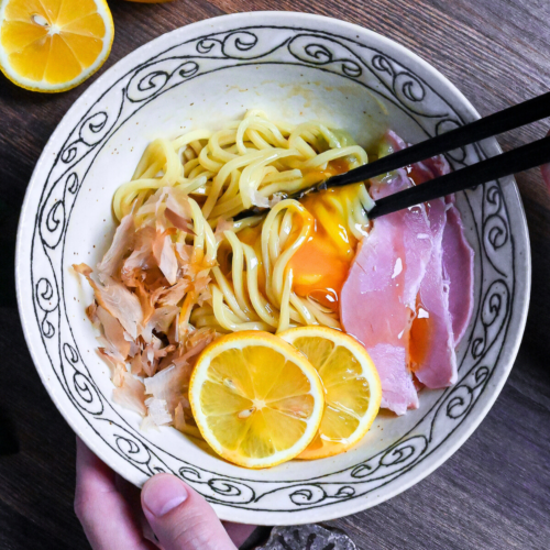 TKM (Tamago Kake Men) topped with bonito flakes, lemon slices, ham, wasabi and a raw egg served in a white bowl with black swirl pattern on rim on a dark wooden surface with lemon in the background