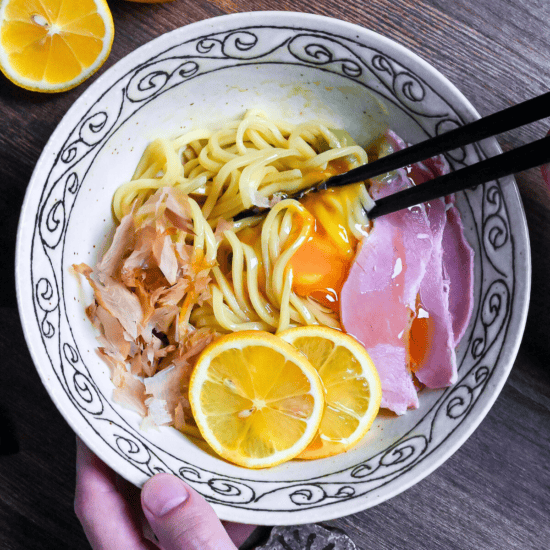 TKM (Tamago Kake Men) topped with bonito flakes, lemon slices, ham, wasabi and a raw egg served in a white bowl with black swirl pattern on rim on a dark wooden surface with lemon in the background