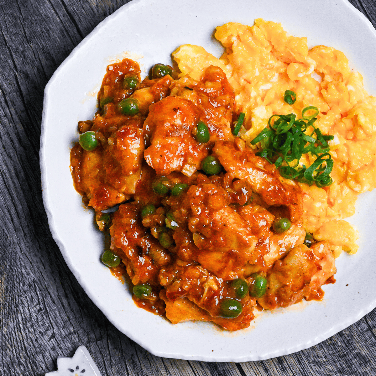 Tori Chili coated with homemade sauce, green peas and buttery scrambled egg on a white plate on a wooden surface top down, thumbnail square