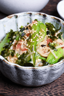 Wasabi-Ae salad with enoki mushrooms and bok choy in a blue and brown bowl on a dark wooden background with umeboshi on a white dish in the background