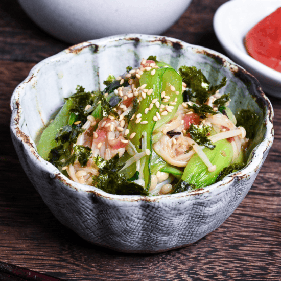 Wasabi-Ae salad with enoki mushrooms and bok choy in a blue and brown bowl on a dark wooden background with umeboshi on a white dish in the background