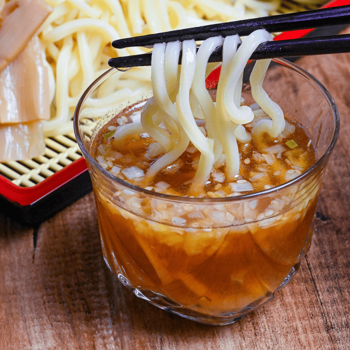 dipping ramen noodles in glass of chilled zaru ramen dipping sauce with cooked and chilled ramen noodles in the background with chashu and boiled eggs